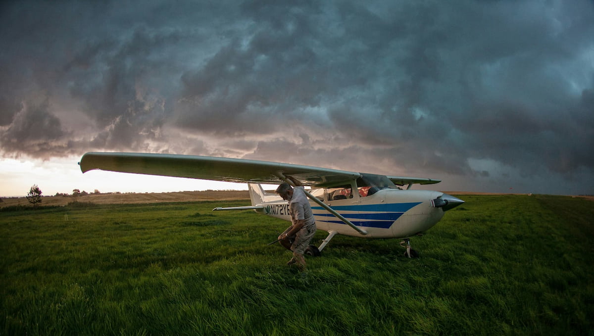 Small airplane on a grass field with a pilot standing nearby under a dramatic sky.