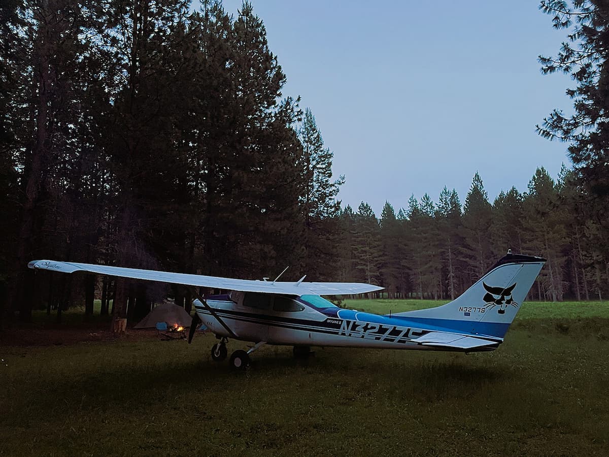 Cessna airplane on a grassy area with trees in the background.  In the isolation of the wilderness is a tent, camp chairs, and a small campfire burning. 