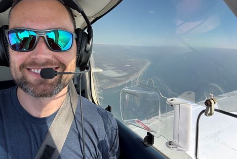 Person wearing sunglasses and a headset inside an airplane cockpit with a clear sky view.