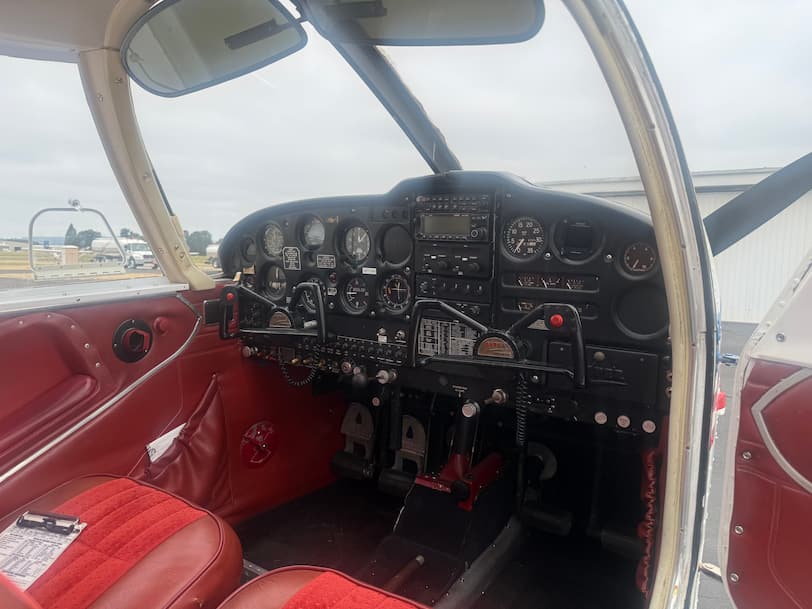 The cockpit interior of a small aircraft, featuring red leather seats, a black instrument panel with multiple analog gauges, and a checklist on the seat.