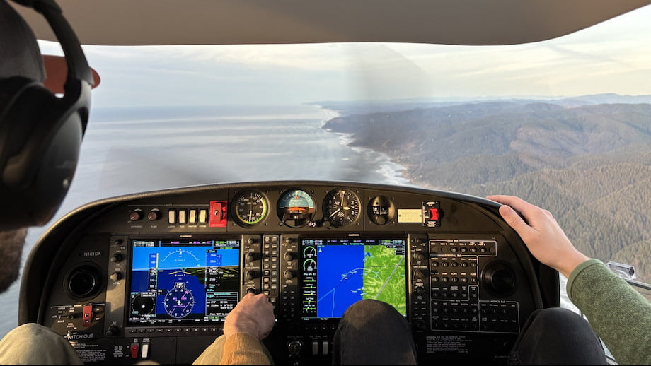 First-person view from inside an DA40 cockpit with a hand on the throttle and another resting on the console, looking out over a coastal landscape with water and forested hills under a bright sky.