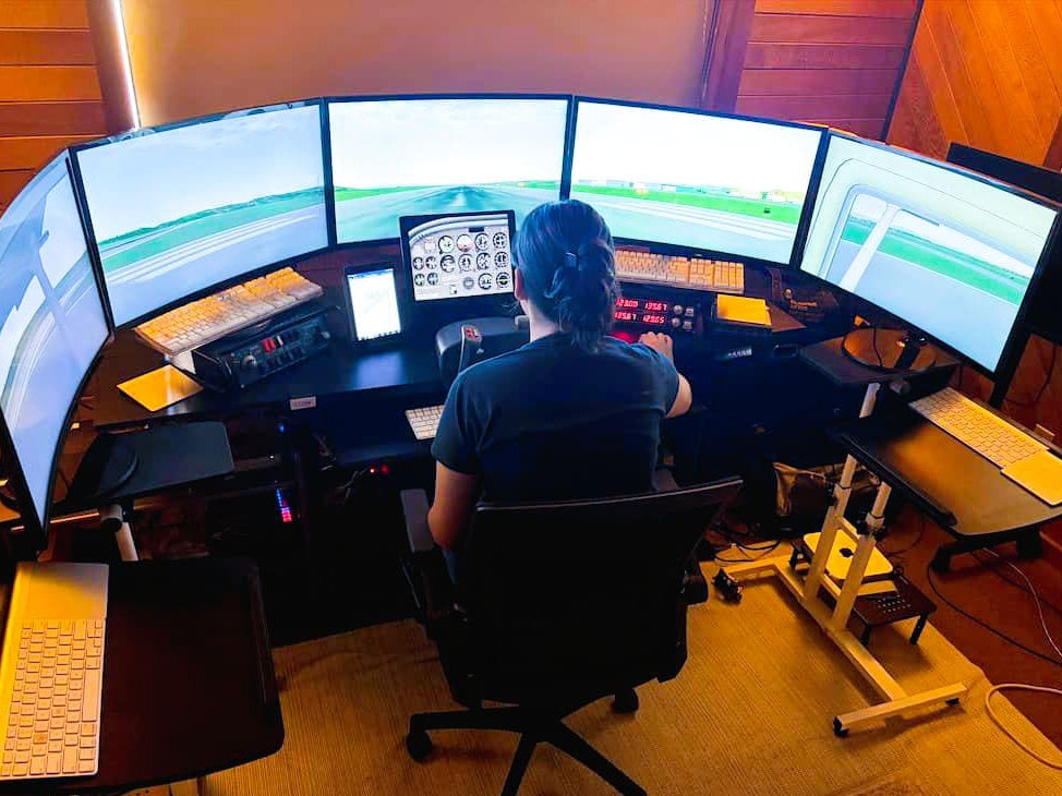A person seated at a curved multi-monitor flight simulation desk, viewed from behind, with cockpit controls and screens visible in a warm wood-toned room.