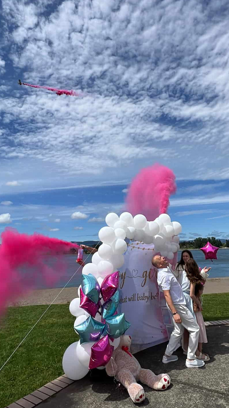 Gender reveal party on river bank park, airplane dropping pink smoke in the background. 