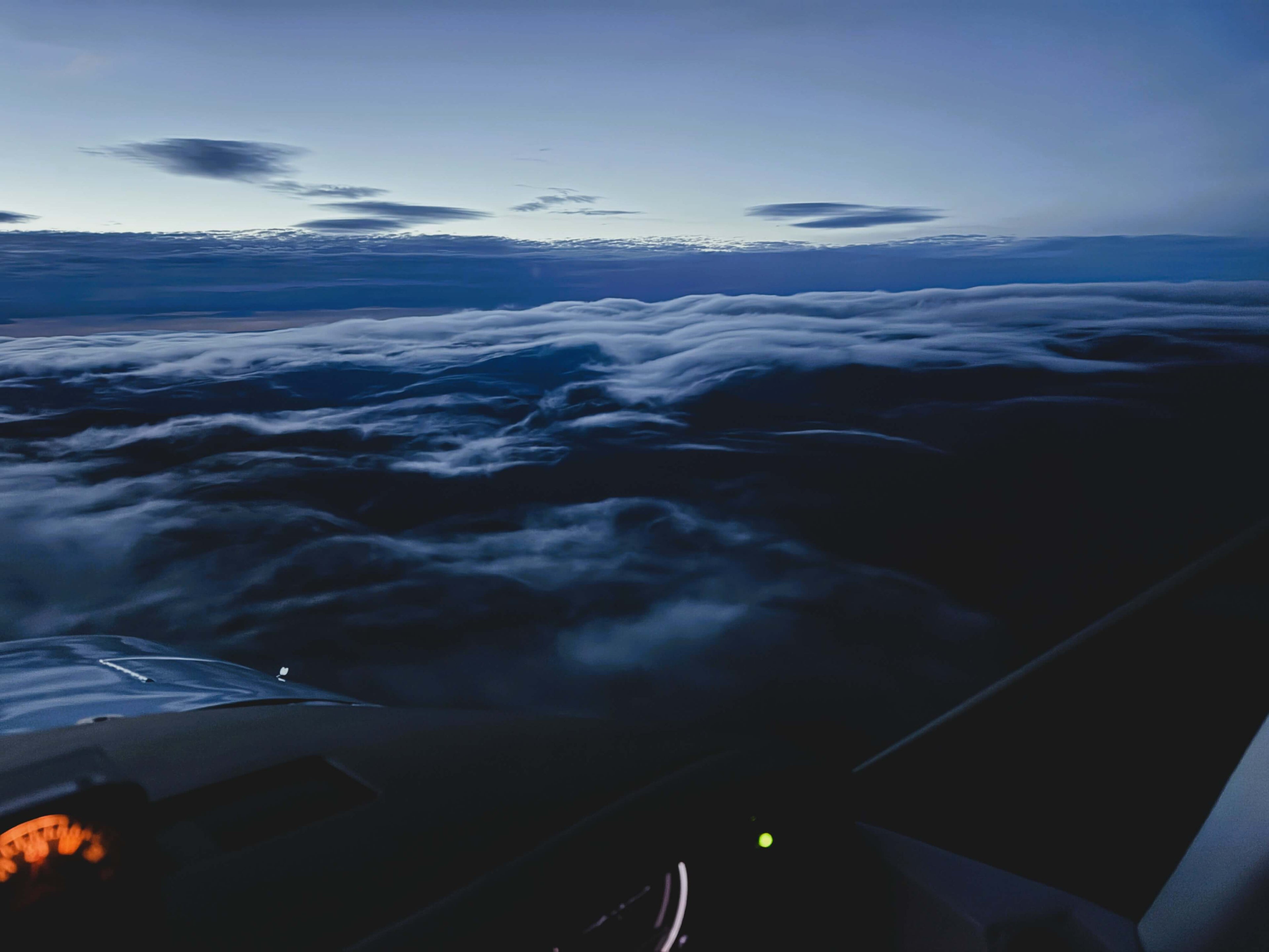 The pilots view from an airplane flying above thick cloud cover that matches the invisible mountainous terrain below. The Airplane cabin is softly lit by instruments and gauges. 