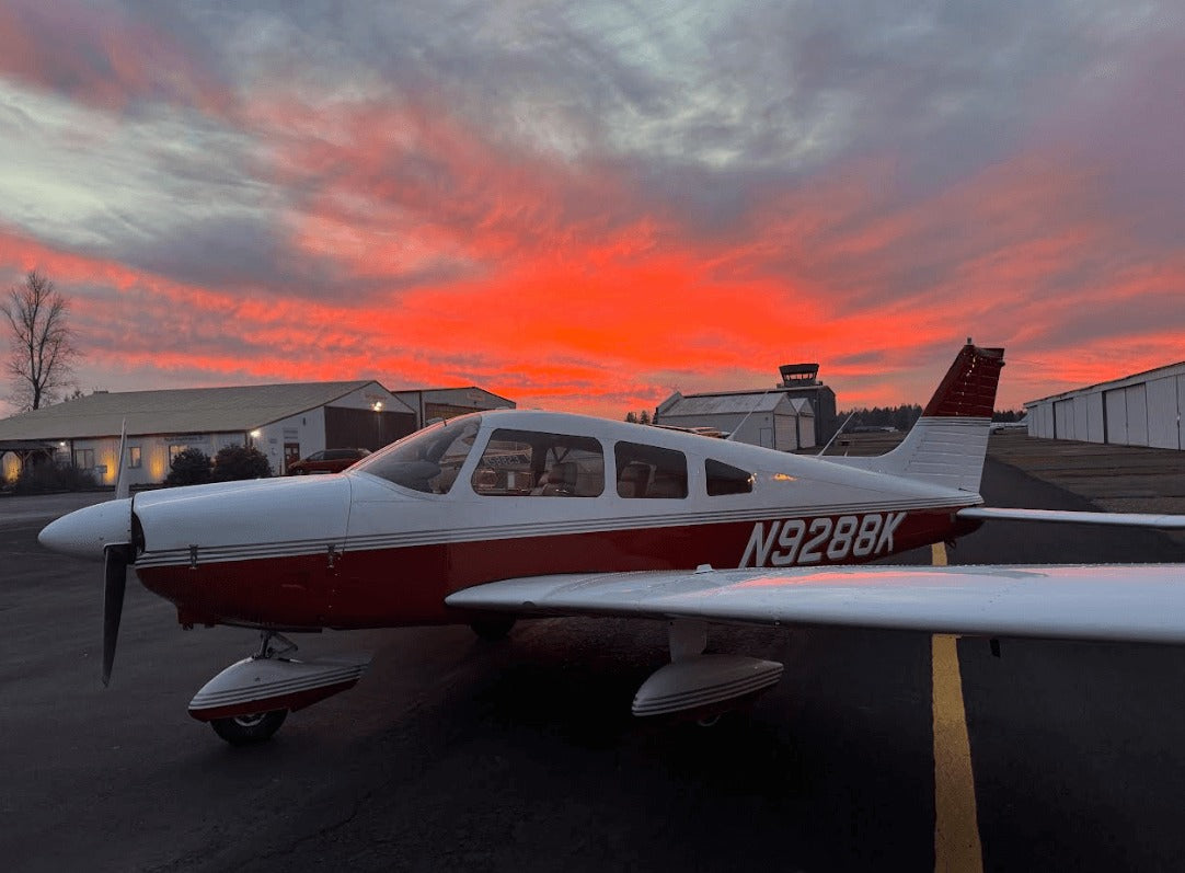 A small white and red single-propeller airplane parked on an airfield tarmac at sunset, with a vivid orange and pink sky, hangars, and an air traffic control tower in the background.
