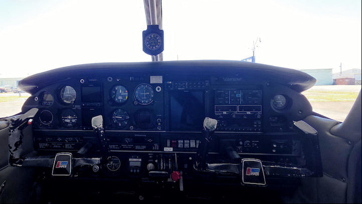 View from inside a small airplane cockpit, showing the instrument panel, dual control, and bright outdoor sky and tarmac in the background.
